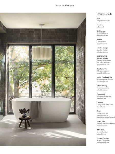 A modern bathroom with a freestanding white bathtub in front of large floor-to-ceiling windows, surrounded by dark tiles. A small wooden stool holds decor beside the tub; trees and greenery are visible outside.