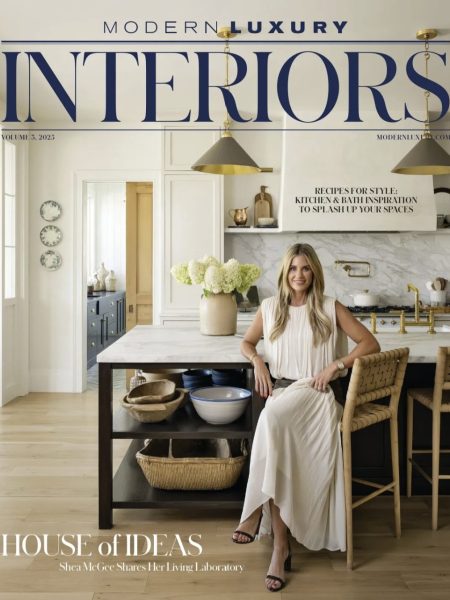 A woman in a white dress sits on a stool in a stylish, modern kitchen with light cabinets, gold accents, and wooden shelves, featured on the cover of Modern Luxury Interiors magazine.
